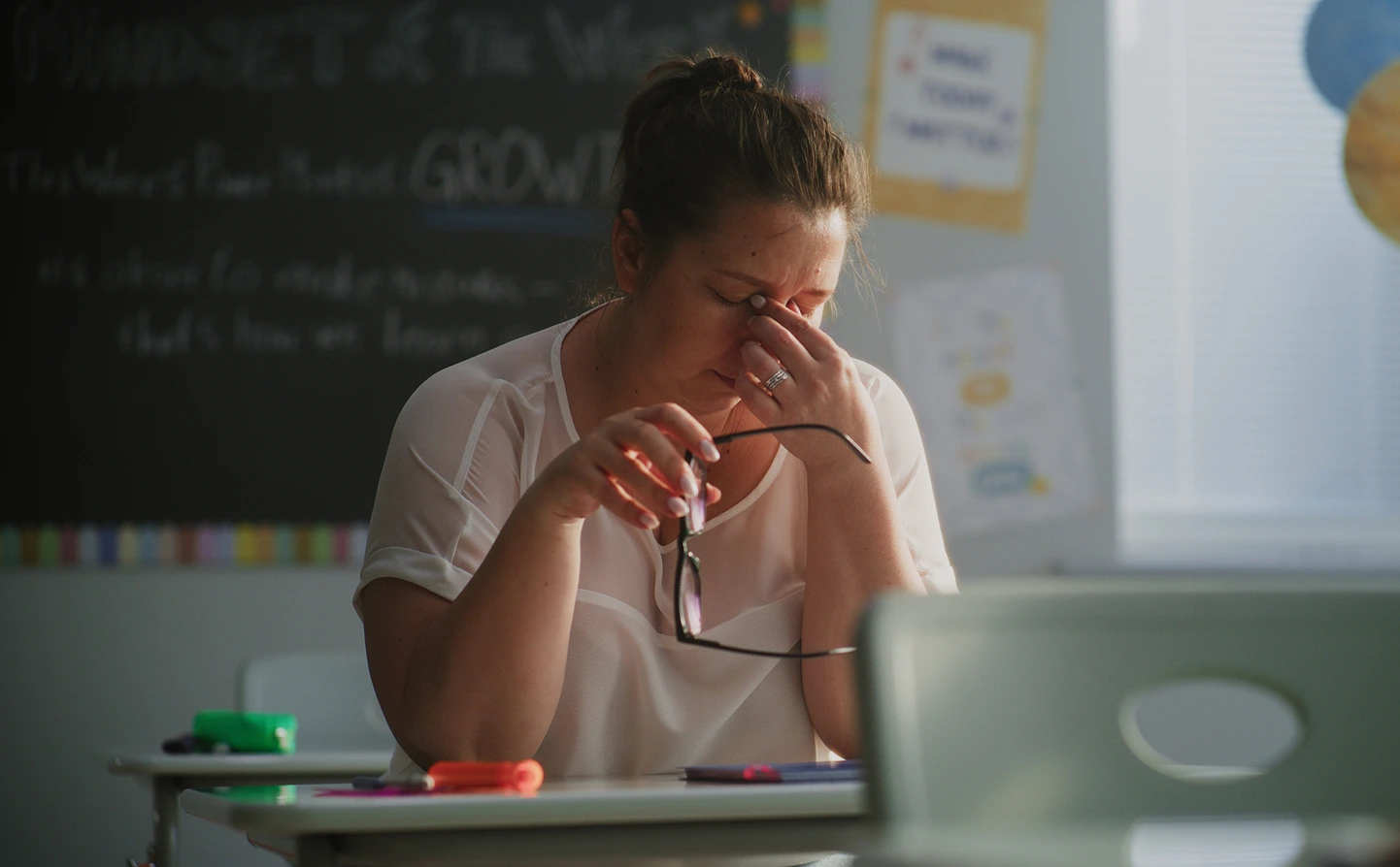 Female teacher in classroom, looking tired. Holding glasses and pinching top of nose with fingers.