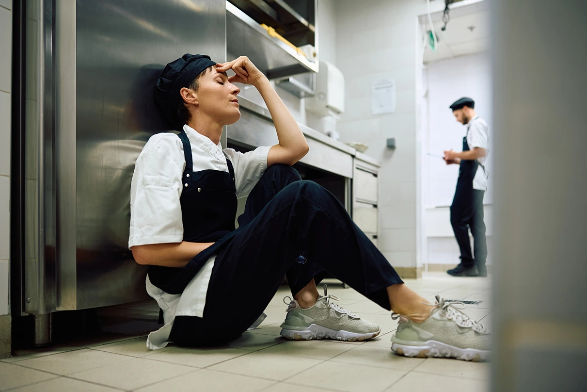 Female chef sitting on the floor of an industrial kitchen with her head in one hand, indicating she is stressed.